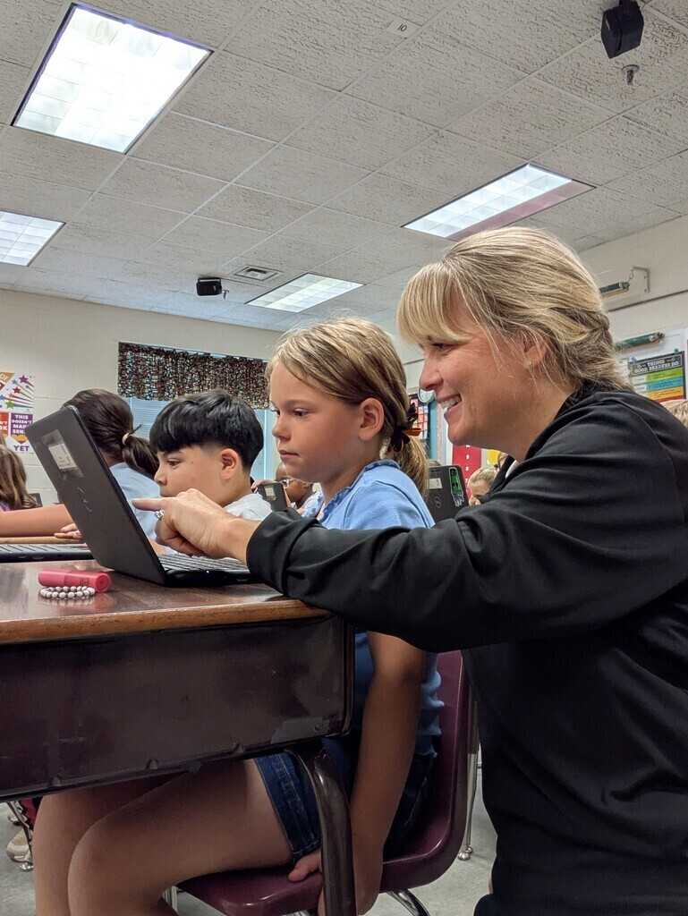 A woman helps a student with a laptop at a desk. Other students sit at desks behind them.