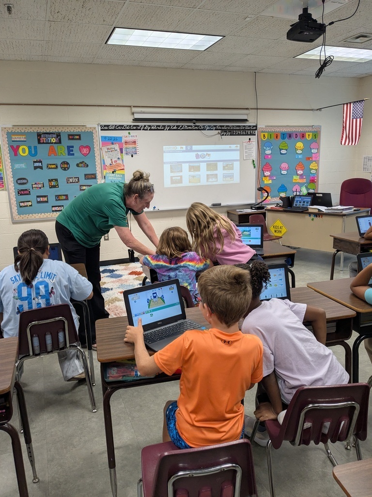 Students sit at desks, facing laptops, in a classroom with a teacher at the front. Brightly colored decorations on the walls.