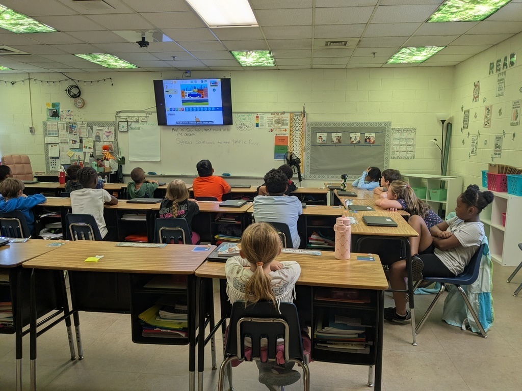 A classroom with a group of students sitting at desks, facing a projector screen. Whiteboard, storage shelves, and a clock are on the walls.