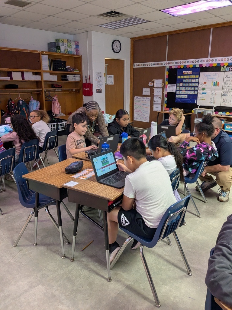 Children seated at desks in a classroom, using laptops. A woman stands behind one of them. Shelves and a bulletin board are in the background.