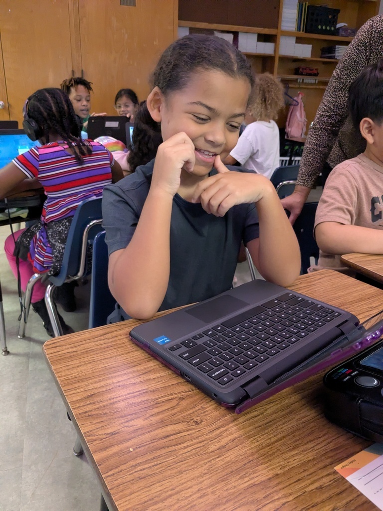 A young girl smiles while working on a laptop at a desk in a classroom.