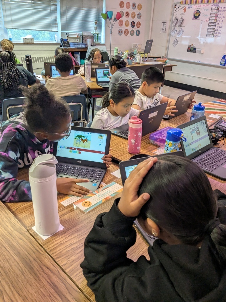 Group of students using laptops at tables in a classroom with a whiteboard and bulletin board in the background.