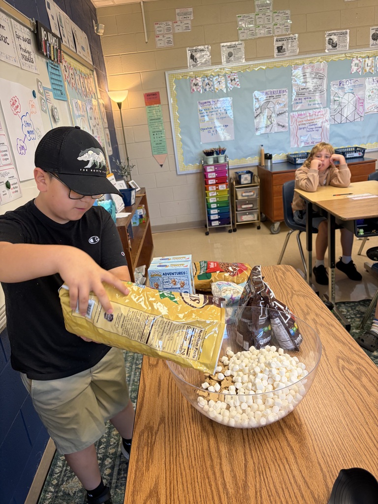 A person wearing a hat pours popcorn into a bowl on a table. Behind them, a classroom has a bulletin board and a student.