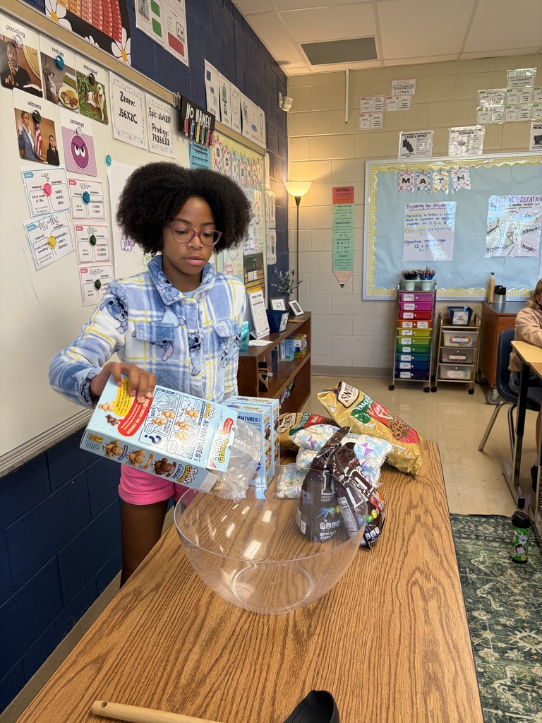 Person standing at a table, pouring cereal into a bowl in a classroom with desks.