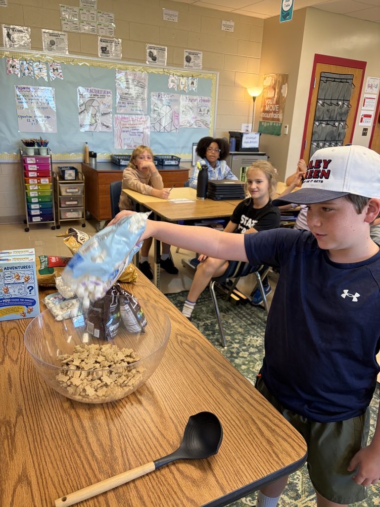 A young boy pouring cereal from a bag into a bowl at a desk. Other students sit at desks behind him.