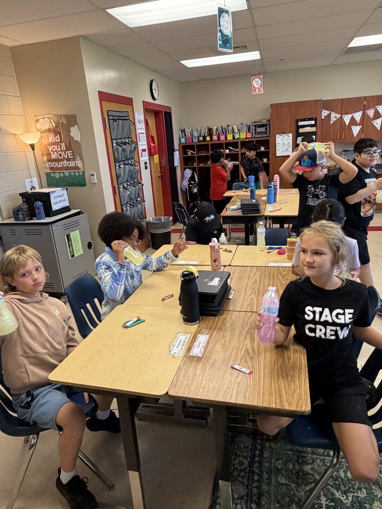Children sitting at tables in a classroom. One child wears a shirt with the word "STAGE" and "CREW".