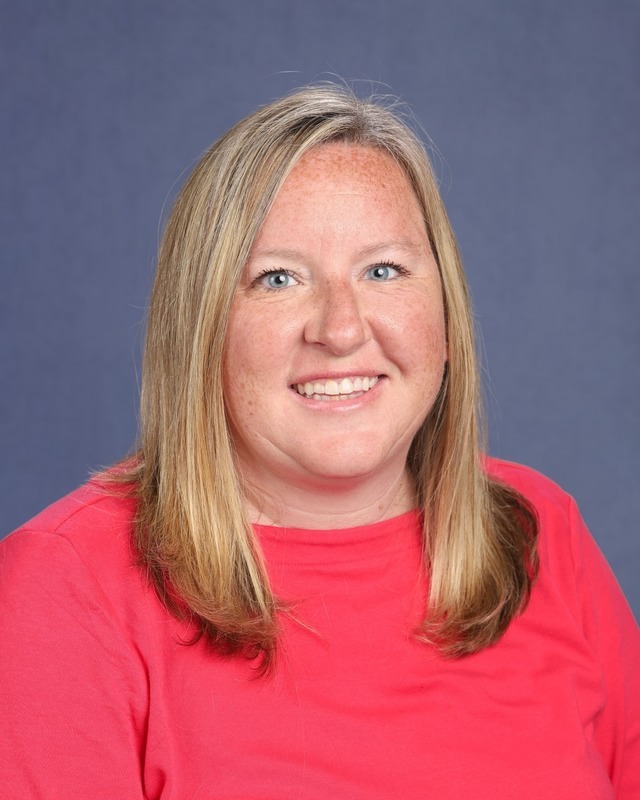 A smiling woman with short blonde hair stands against a blue background. She wears a pink t-shirt and looks forward.