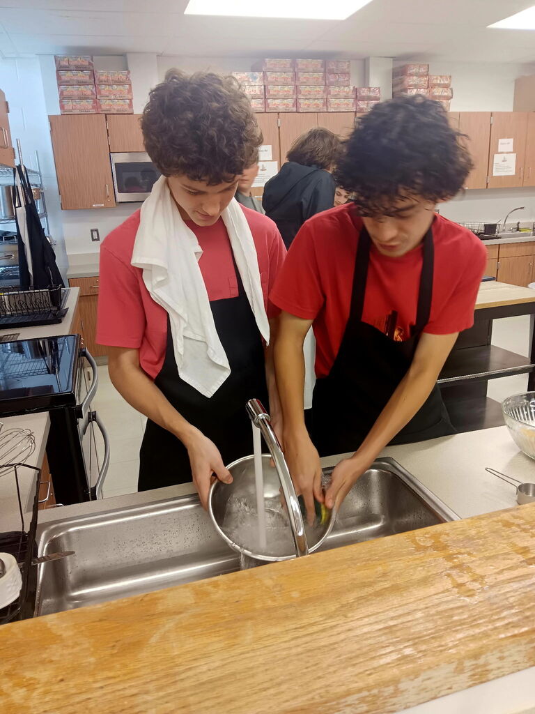 Two individuals in red shirts and aprons washing dishes at a sink in a kitchen.