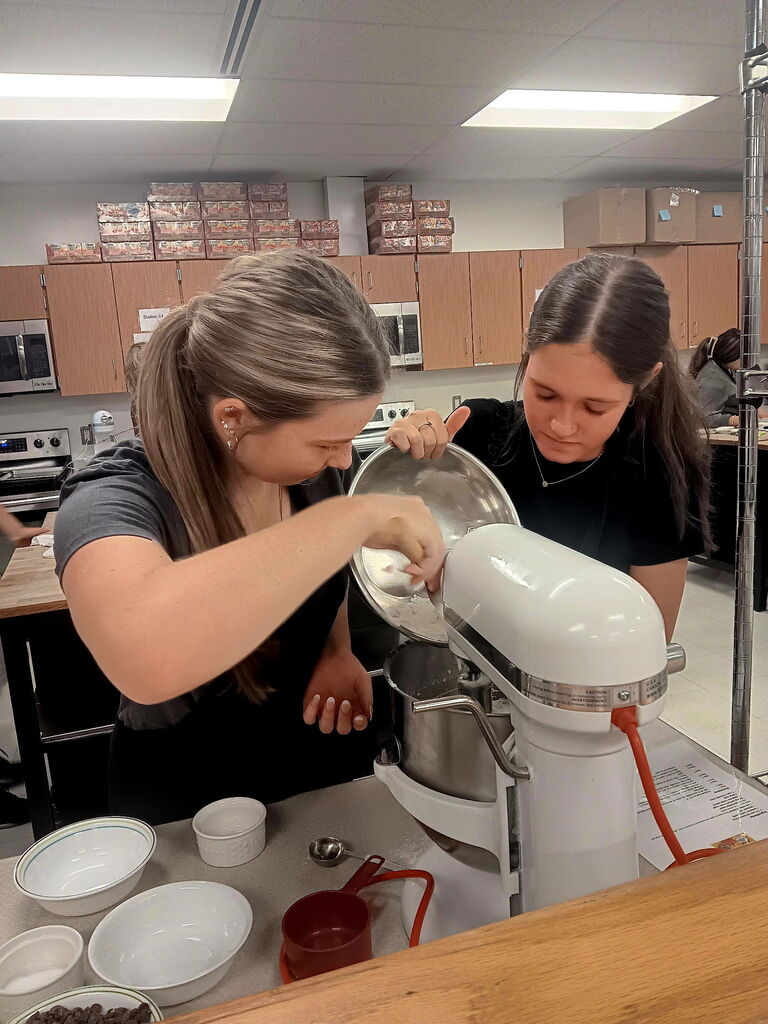 Two women prepare ingredients for a recipe using a large mixer, with several bowls and measuring cups on a kitchen counter.