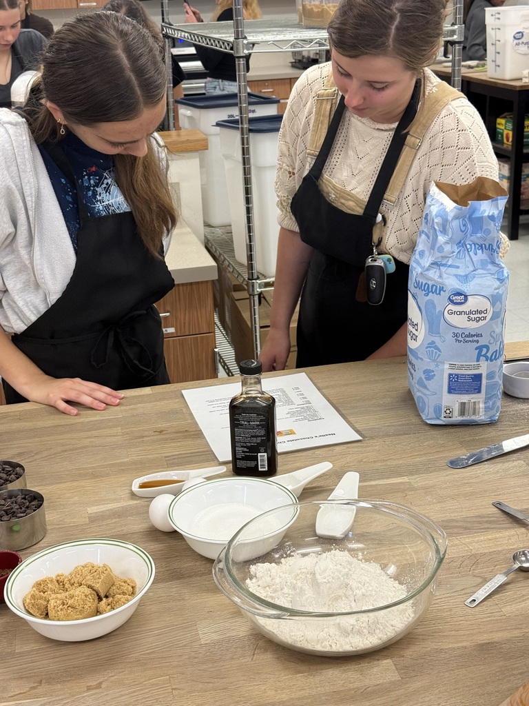 Two women wearing aprons in a kitchen with ingredients and utensils on a table, including a bowl of flour.