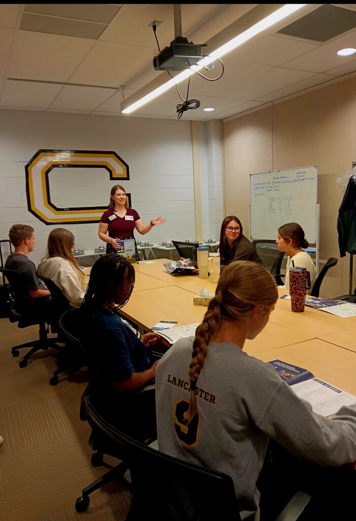 A group of students seated at a long table, a woman stands at the front of the room.