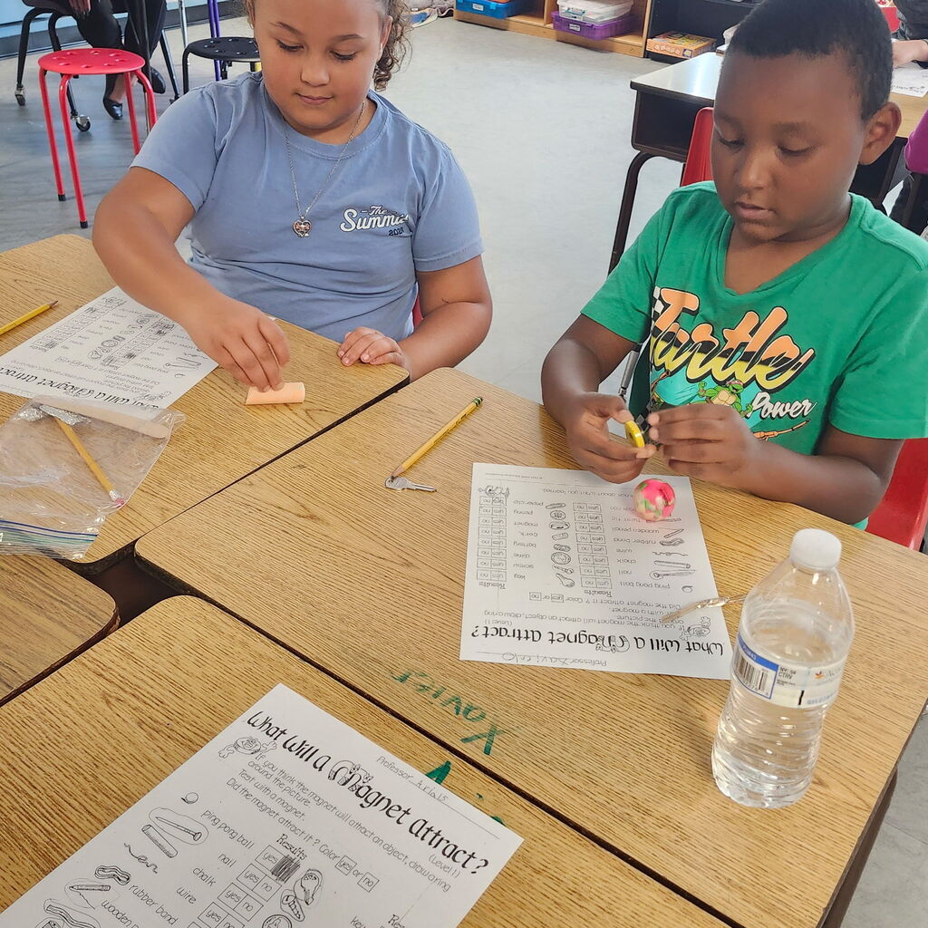 Two children seated at a desk, engaged in a school activity with printed sheets and objects.