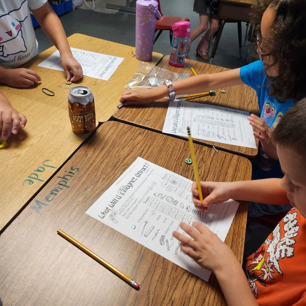 Four students work on a table. A boy in orange writes on paper. Next to him, a girl in blue holds a pencil. Behind them, a girl in white works at a table.