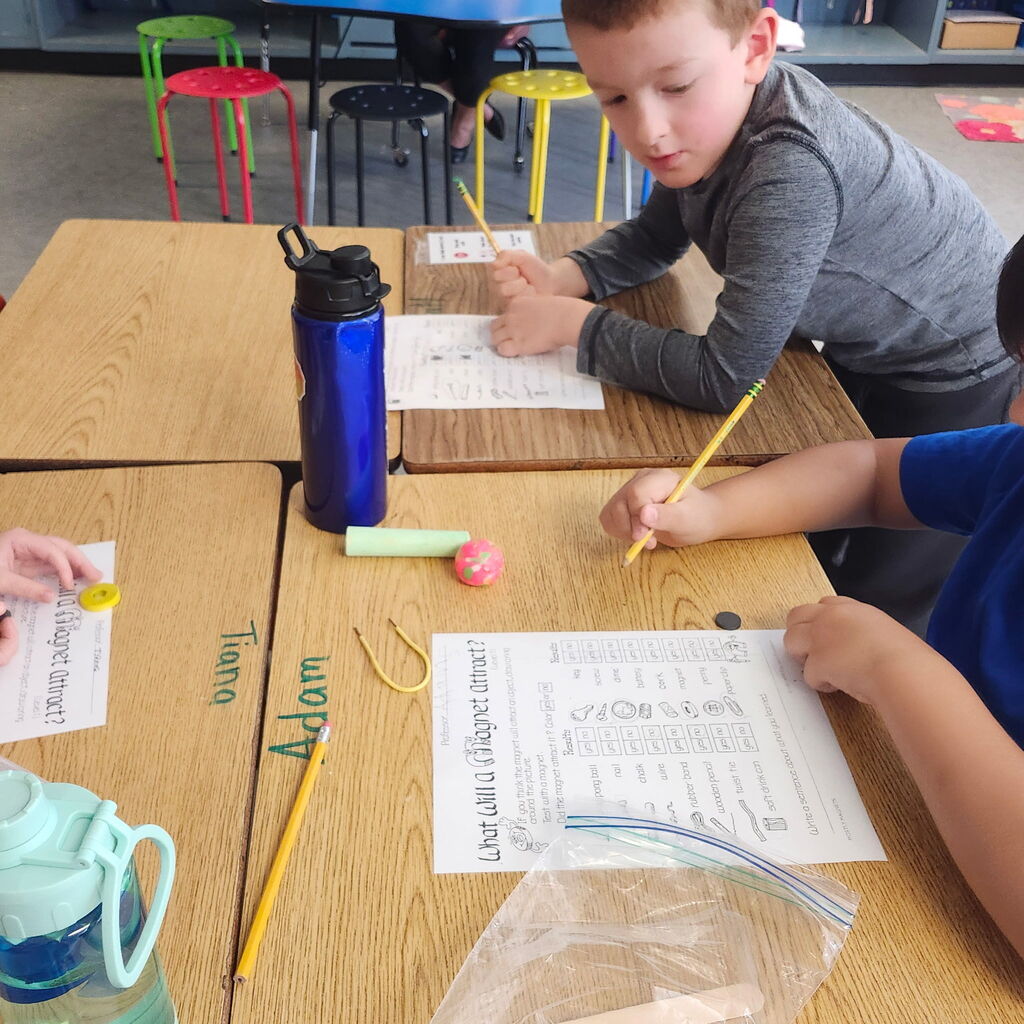 Two children seated at desks in a classroom, writing on papers. One holds a pencil, the other a pen.