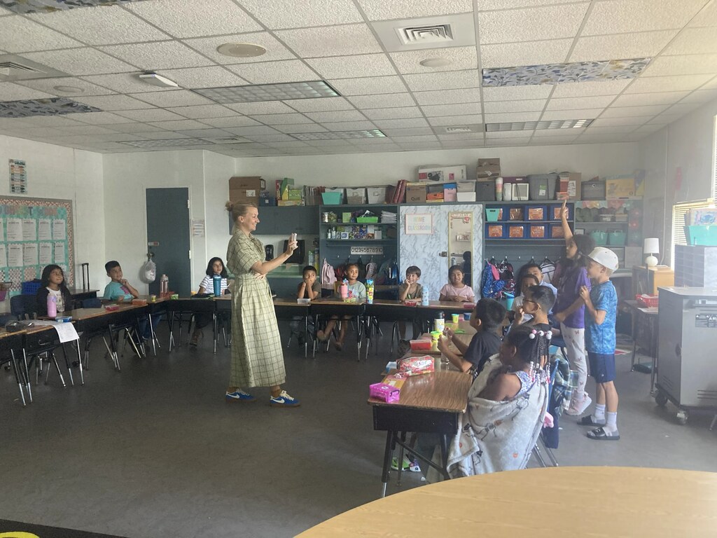 A classroom with rows of desks, students seated, and a woman standing in front of them, holding a microphone.