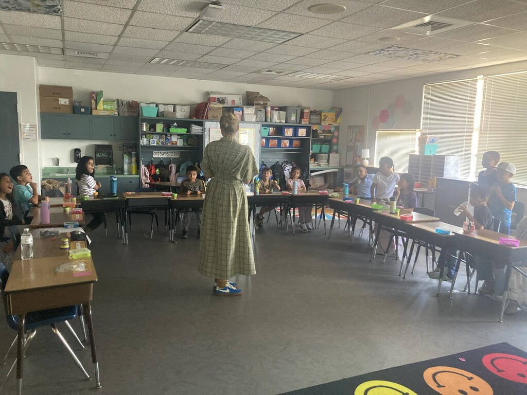 A classroom with a teacher standing in front of children seated at long tables.
