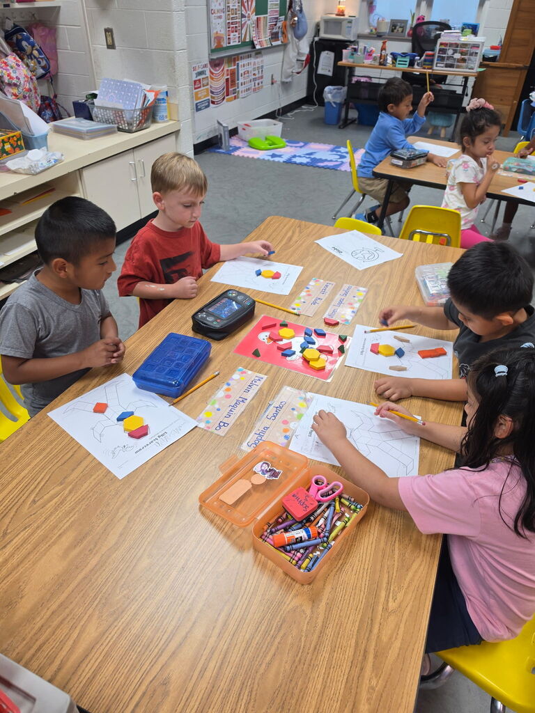 Five children work at a table, using colored pencils and geometric shapes. Other children sit at tables behind them.