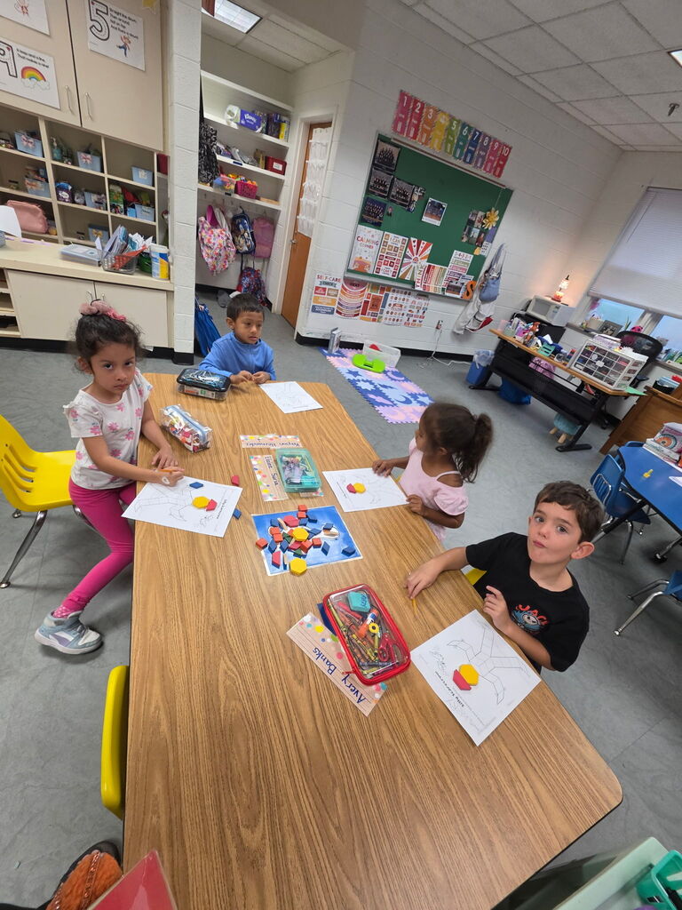 Four young children sitting at a table with educational materials, in a classroom with bright lighting and shelves of books.