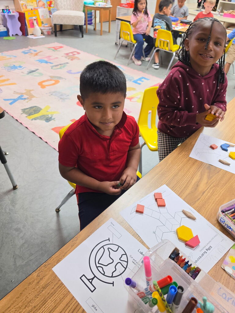 Two young children smile at the camera, one standing and the other seated, surrounded by colorful shapes and markers on a classroom table.