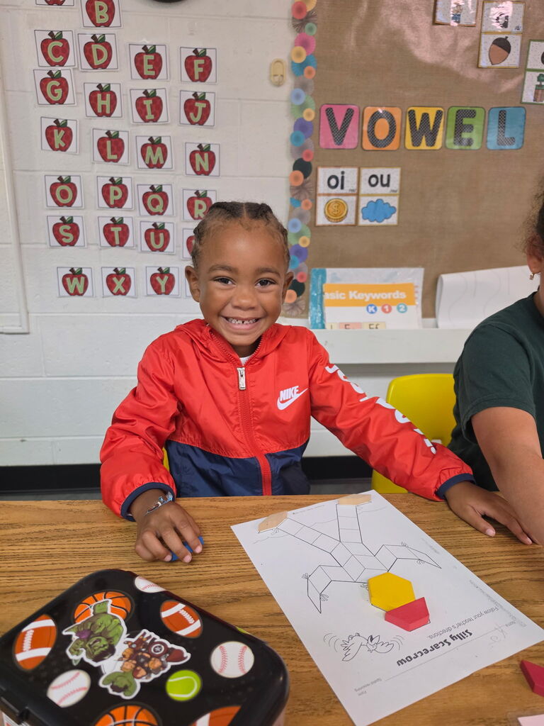 A child in a red jacket smiles while sitting at a desk with a paper and lunchbox.