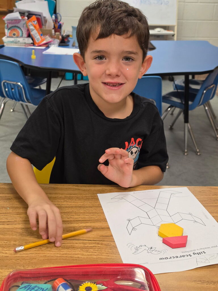 A child sitting at a desk in a classroom, holding a pencil and looking at a drawing.
