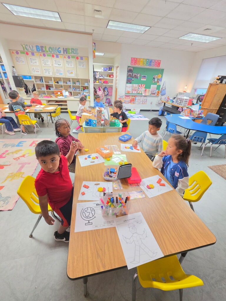 Classroom with tables, chairs, and children seated. A table has papers and markers. Other tables, chairs, and posters are in the background.