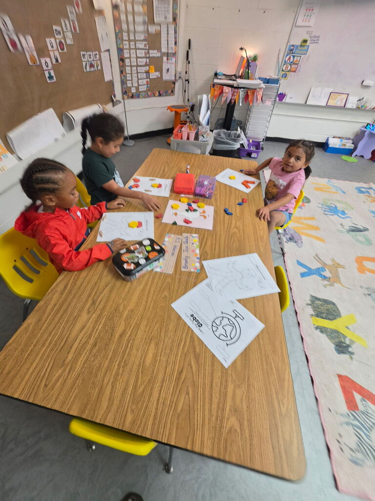 Three young children seated at a table, engaged in an activity with papers, crayons, and a tray.