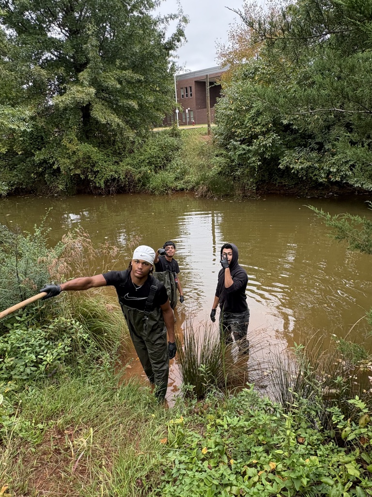Three people in dark clothing, gloves, and hats wade in a brown river, surrounded by trees.