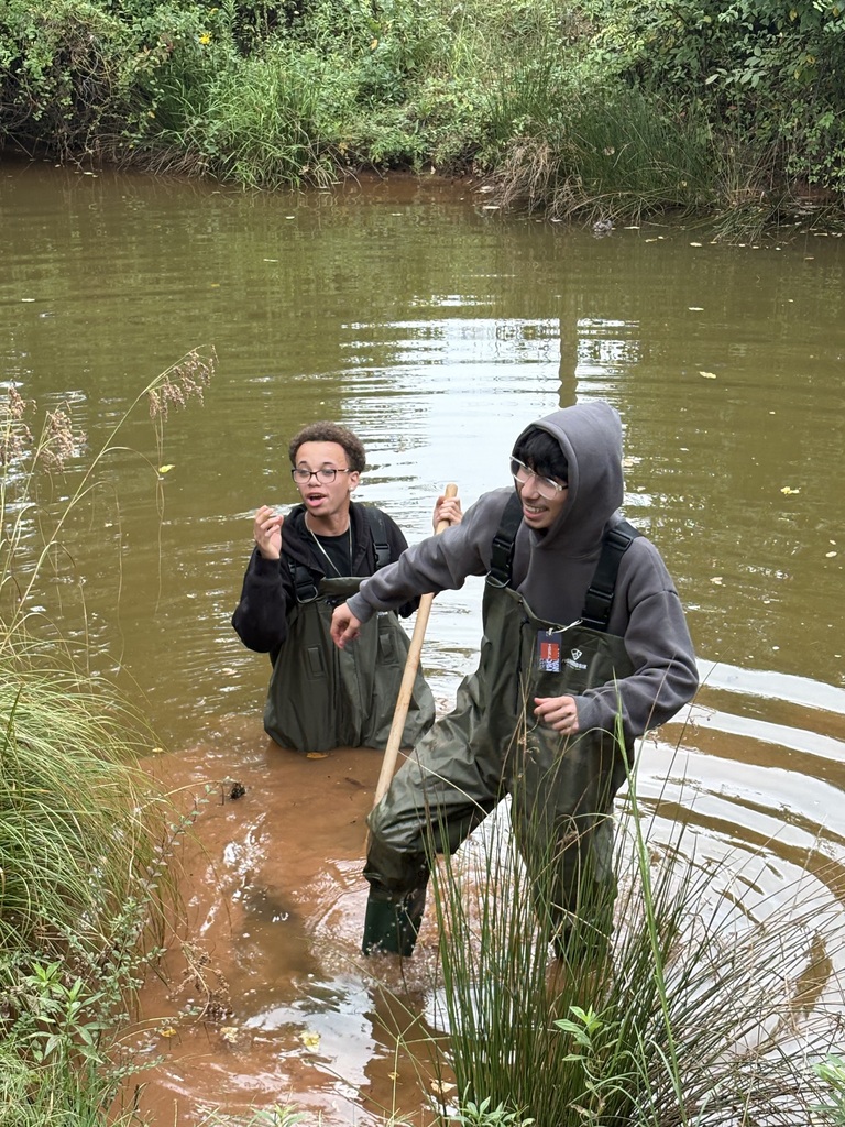 Two people in rain gear stand in shallow water, holding sticks, and looking at the ground.
