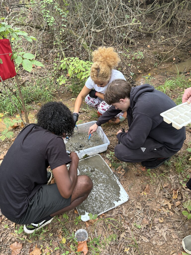 Three people examine a container of water outside. One is kneeling and two are seated. Greenery and fallen leaves surround them.