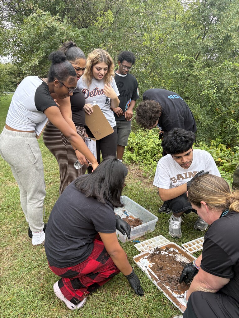A group of students outdoors, with some bent over examining soil. One student holds a tray. Trees and greenery in background.