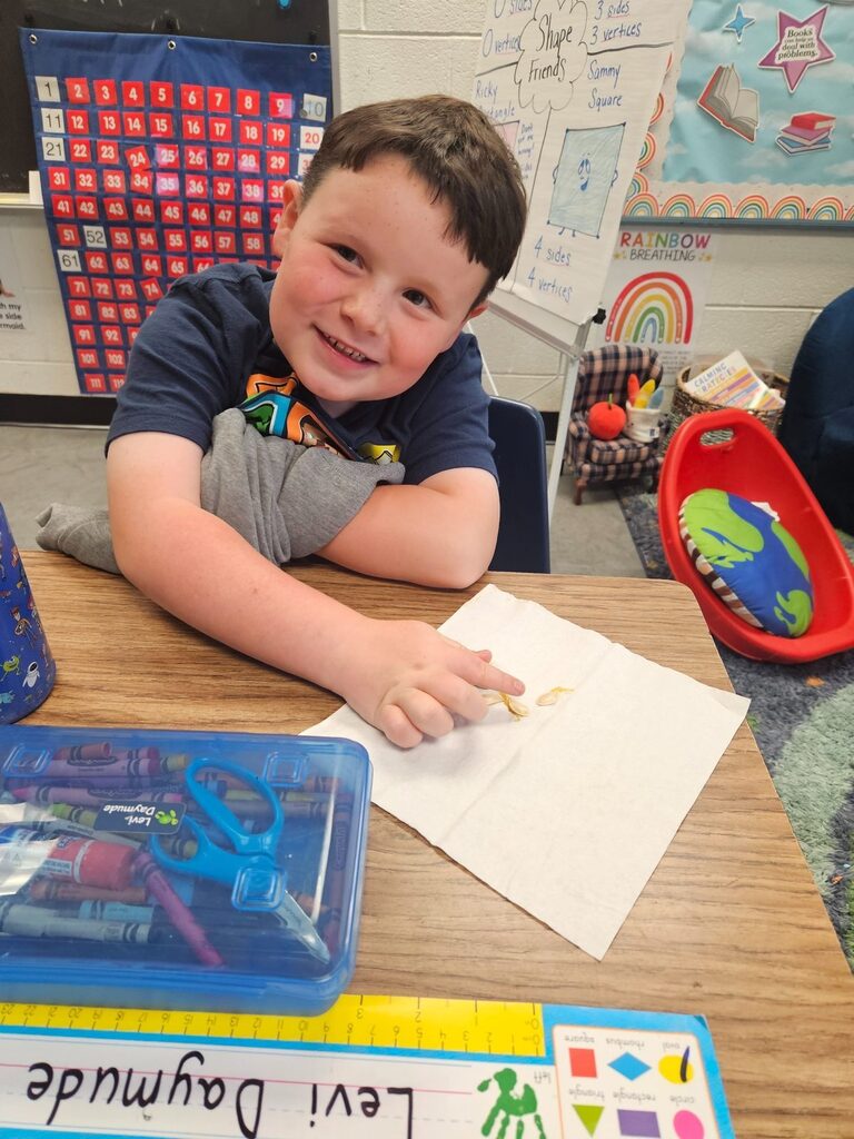 A young boy in a blue shirt smiles as he works on a craft project at a desk.