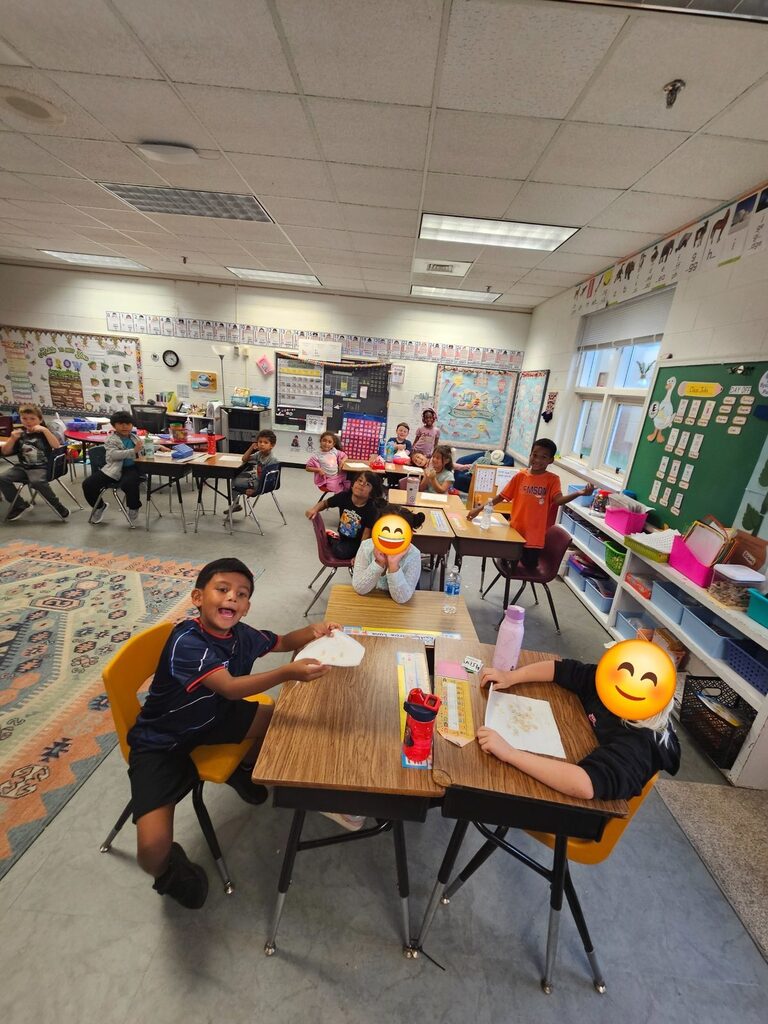 A classroom with children at desks. Desks have books and cups on them. Faces have emojis over them.