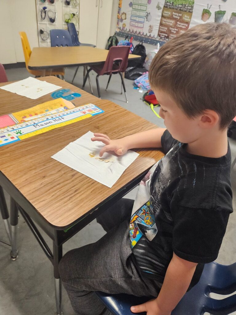 Child at a desk with hands on a white piece of paper in a classroom.