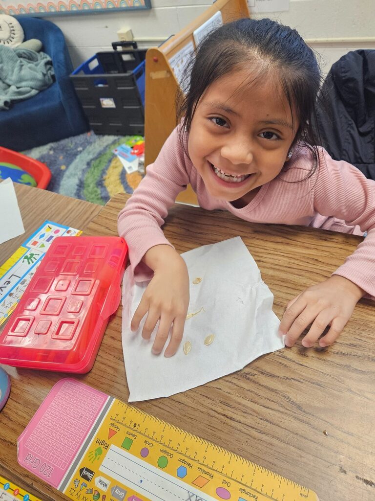 Child at desk, smiling, holding a piece of paper with yellow objects. Behind, a storage box and chair.
