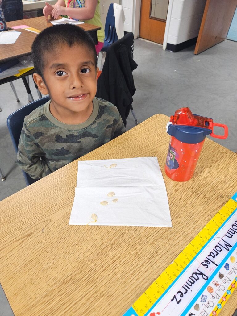 A child at a desk with a ruler, a cup, and a paper with coins. Background shows classroom with other students.