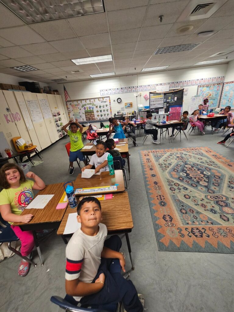 A classroom with desks. Students seated and one raising hand. Brightly lit room with a rug on the floor.