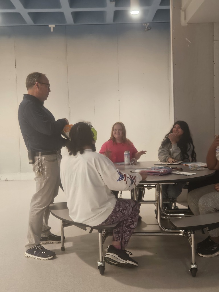 A staff member stands beside a cafeteria table speaking with a group of CMS students. Several students smile and laugh as they listen and interact during the activity.