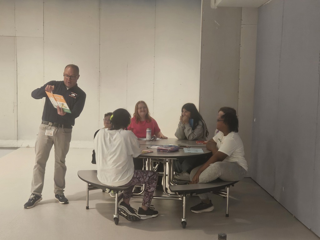 A staff member demonstrates or explains an item to students gathered around a cafeteria table. Students look on with smiles and interest during the group activity.