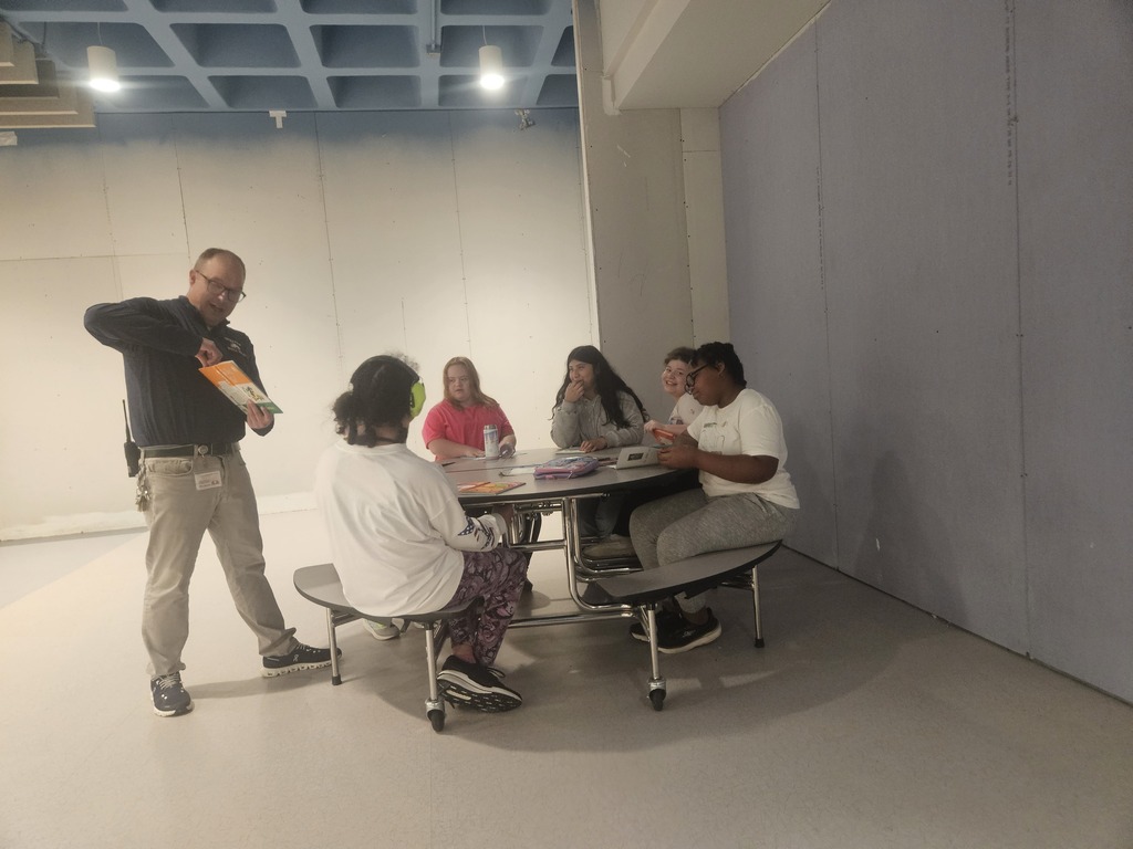 A staff member holds up an item while talking with a group of students seated around a cafeteria table. The students watch, smile, and participate in the conversation.