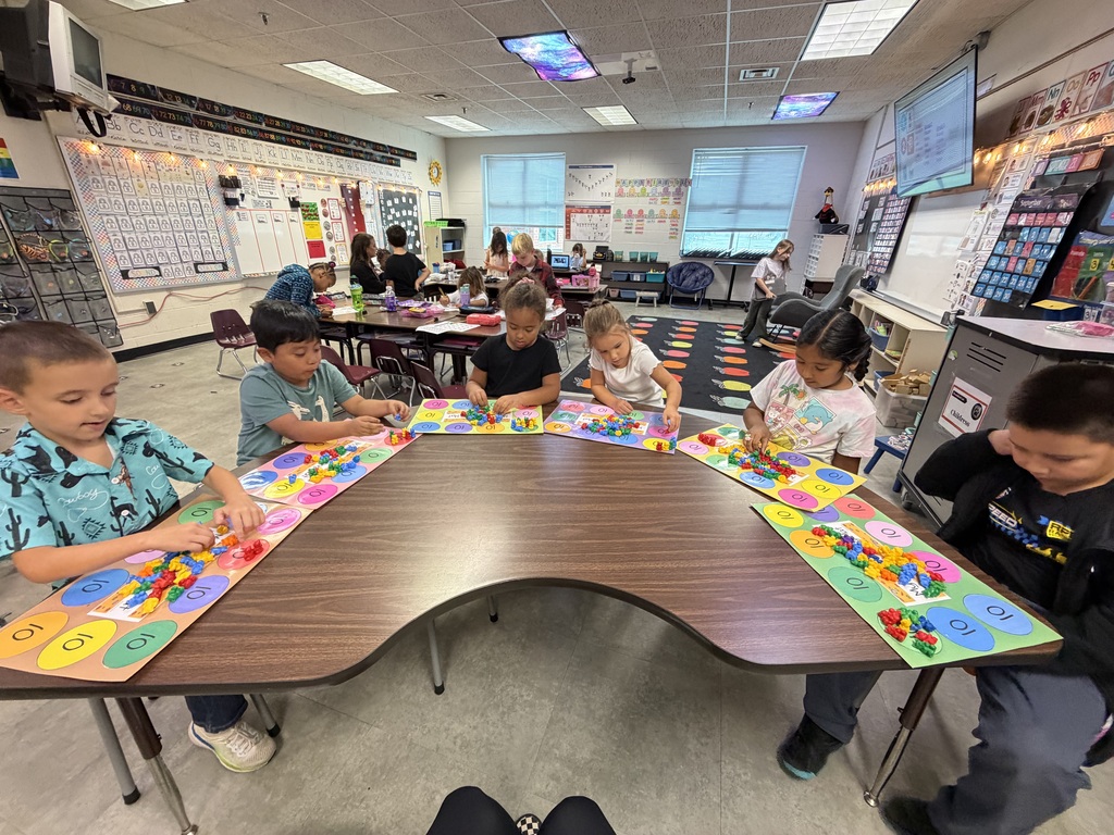 Kids sit at a table, engaging with colorful blocks. Bright lights and a whiteboard are in the background.