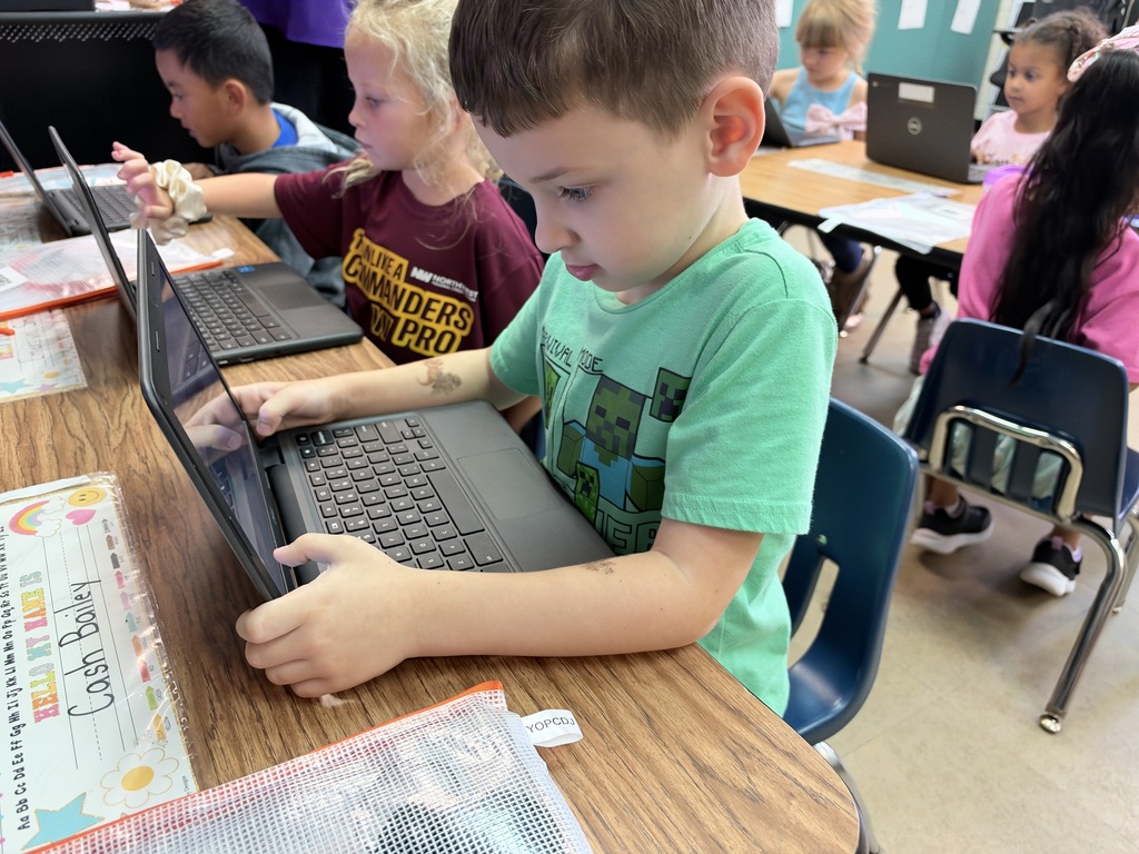Several children using laptops in a classroom. One child, wearing a green shirt, focuses on a laptop.