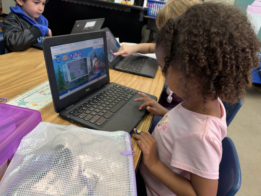 A child uses a laptop with a colorful game screen on it. A boy sits behind her.
