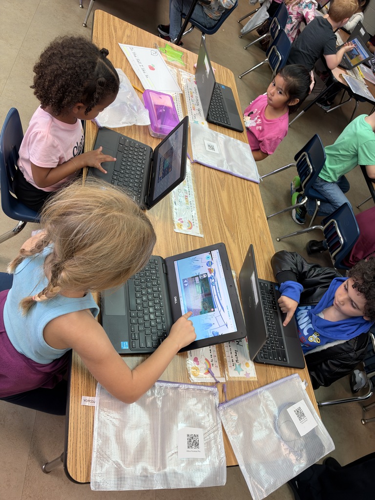 Children seated at a table, using laptops, with one person in a wheelchair. Some students sit on chairs, and some on the floor.