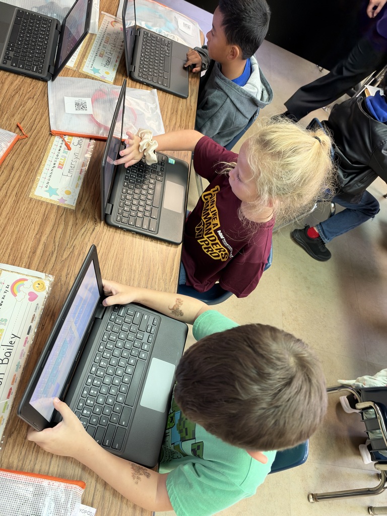 Kids using laptops on a table. One child uses a laptop with a yellow lanyard.
