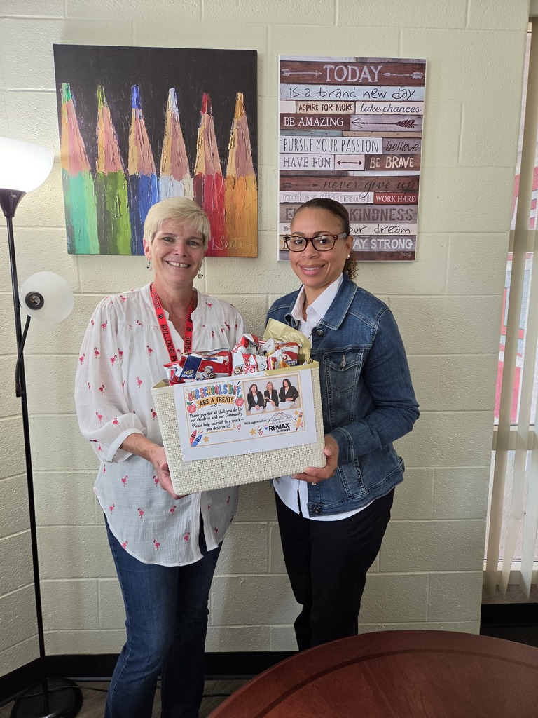 Two women stand, one holding a box, in a room with a colorful pencil poster and a window.
