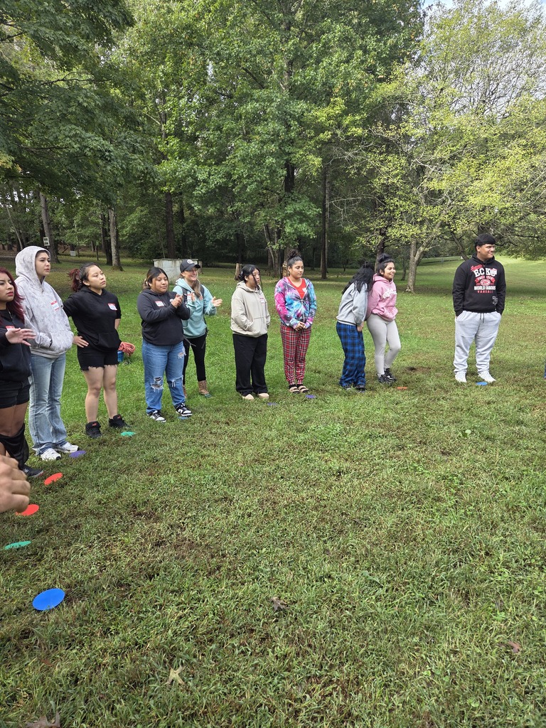 Nine people in various tops and pants gather in a grassy field with trees in the background.