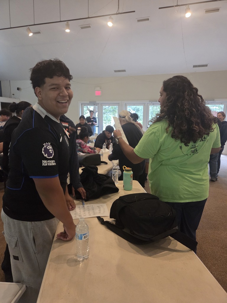 A man and a woman stand at a table in a large room. The man smiles at the woman.