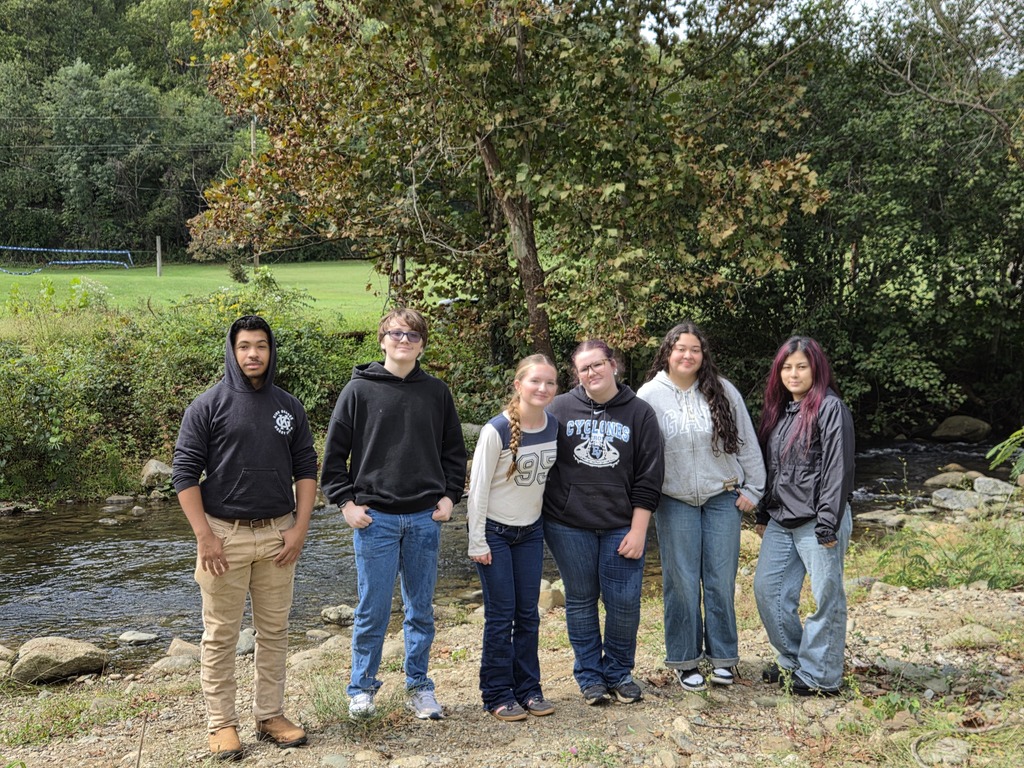 Six individuals, in casual attire, stand by a small stream with greenery in the background.