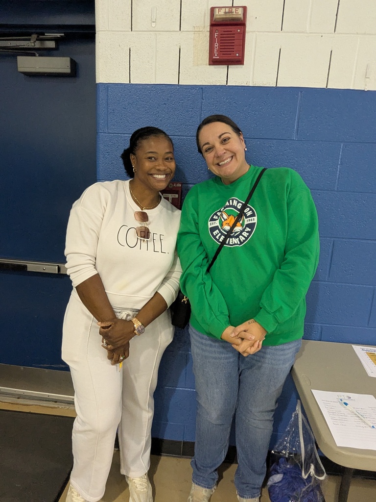 Two women, one in white and one in green, stand smiling next to a blue wall. One has a coffee-themed shirt, and they are near a table.